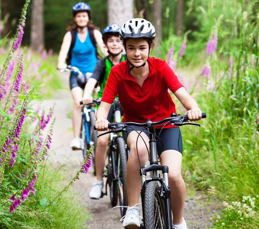 Image of mother and two sons riding their bikes down trail lined with purple flowers.