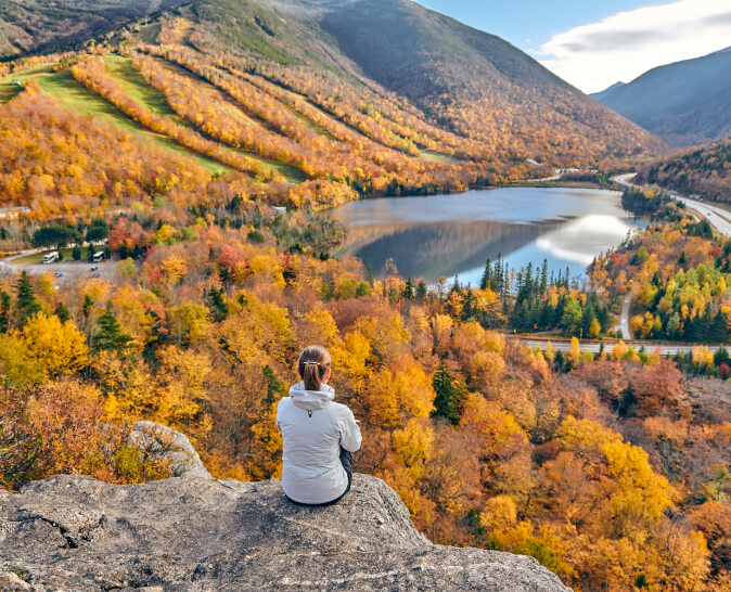 Image of lady sitting on a bluff looking at a lake and fall trees.